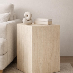 Könnte beinhalten: Ein hellbeige, rechteckiger Beistelltisch mit strukturierter Oberfläche. Eine weiße, abstrakte Skulptur und zwei gestapelte Bücher stehen oben. Der Tisch steht neben einem beigen Sofa auf einem neutralen Teppich.
