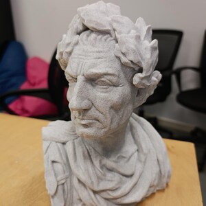May include: A light grey bust of a person with a stern facial expression, wearing a laurel wreath on their head and draped fabric over their shoulders. The sculpture has a textured, stone-like finish and is depicted from the chest up.