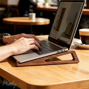 Könnte beinhalten: Ein Laptop-Computer liegt auf einem Holzständer, der für eine komfortable Nutzung angewinkelt ist. Der Ständer hat eine dunkelbraune Farbe, und der Laptop-Bildschirm zeigt eine Website an. Eine Tasse Kaffee steht in der Nähe auf einer Untertasse. Das Wort "Plushy" steht unten links.