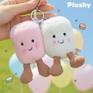 Könnte beinhalten: Zwei Plüsch-Schlüsselanhänger in Form von Eis am Stiel. Einer ist rosa, der andere weiß, beide mit lächelnden Gesichtern, rosigen Wangen und braunen Füßen. Das Wort "Plushy" steht oben rechts. Die Schlüsselanhänger sind an einem silbernen Schlüsselring befestigt.