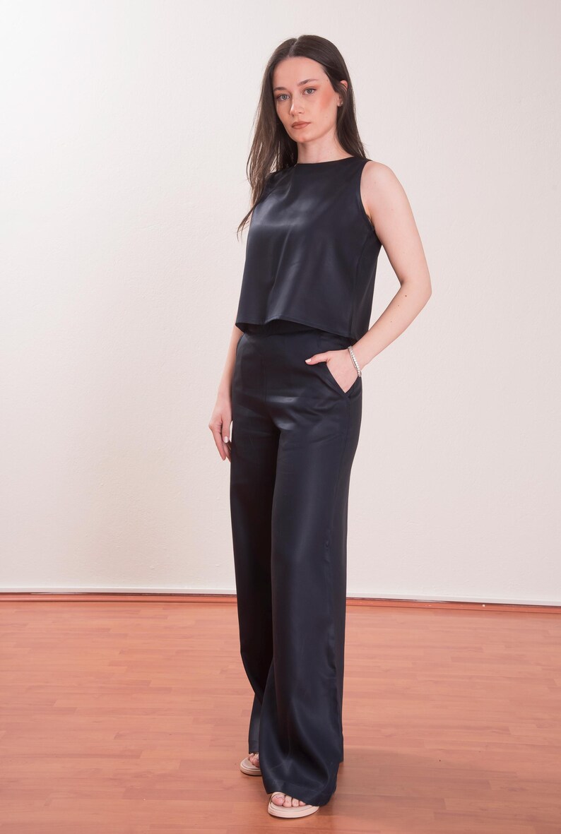 May include: A sleeveless, navy blue top and matching wide-leg pants. The top has a round neckline and a cropped design. The pants feature a high waist and pockets. The outfit is made of a smooth, shiny fabric, creating a sleek, elegant look.