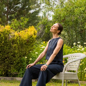 May include: A woman wearing a navy blue sleeveless top and matching wide-leg pants sits on a white wicker chair in a garden. She wears black sandals and a necklace. The background features green foliage and yellow flowers.