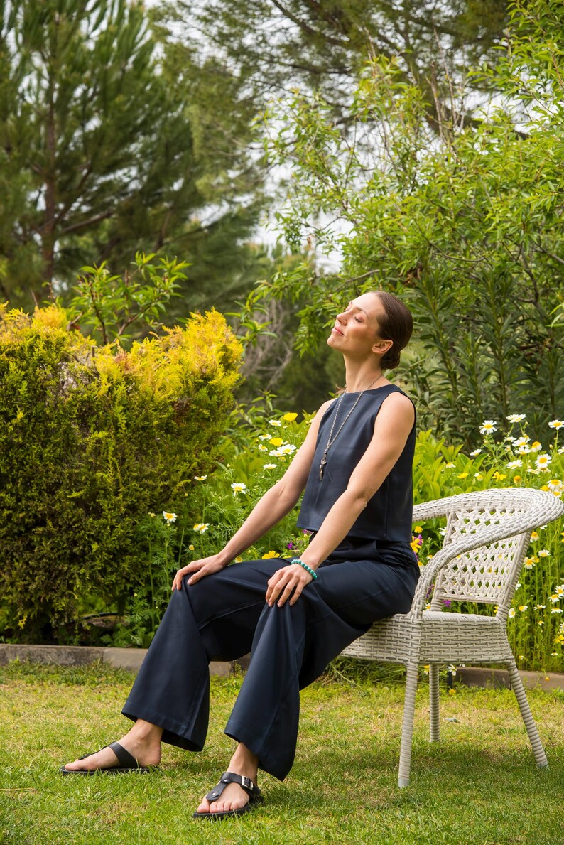 May include: A woman wearing a navy blue sleeveless top and matching wide-leg pants sits on a white wicker chair in a garden. She wears black sandals and a necklace. The background features green foliage and yellow flowers.