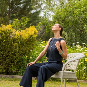 May include: A person wearing a navy blue sleeveless top and wide-leg pants, seated on a white wicker chair in a garden. The person is wearing black sandals and a necklace. The background features green foliage and yellow flowers.