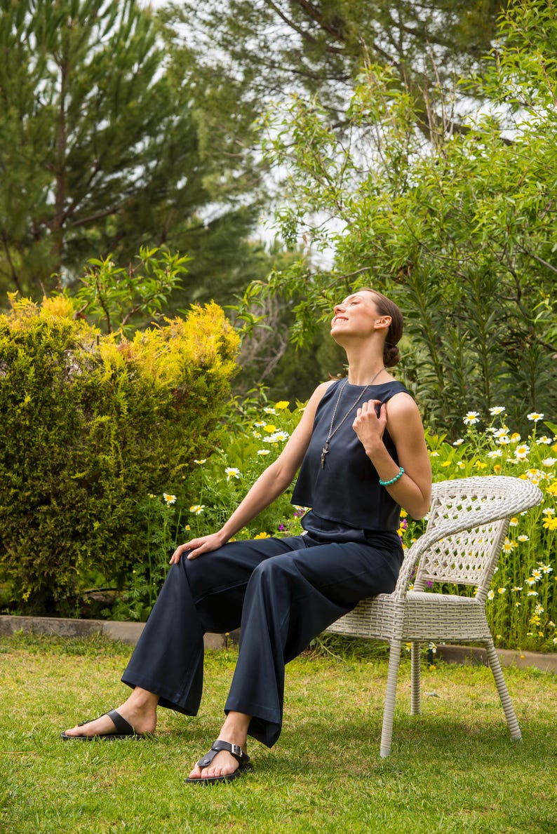 May include: A person wearing a navy blue sleeveless top and wide-leg pants, seated on a white wicker chair in a garden. The person is wearing black sandals and a necklace. The background features green foliage and yellow flowers.