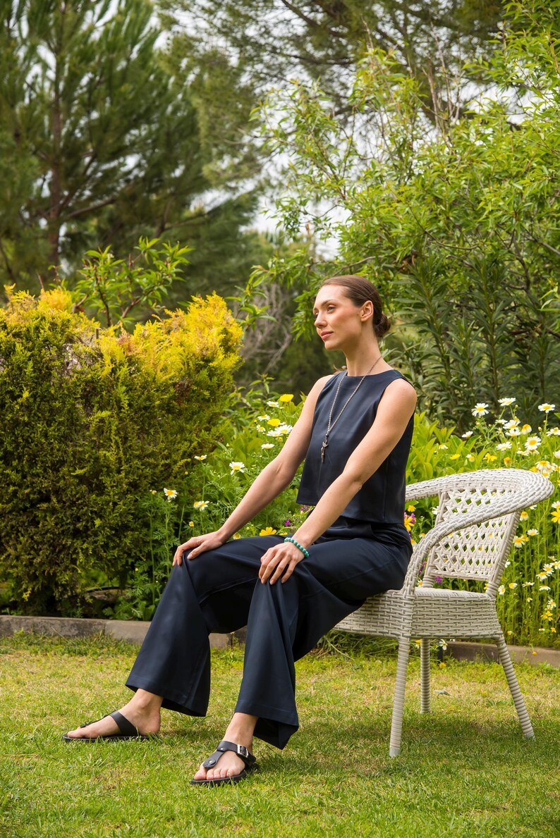 May include: A woman wearing a navy blue sleeveless top and matching wide-leg pants, seated on a white wicker chair in a garden setting. She wears black sandals and a necklace. The background features green foliage and flowers.