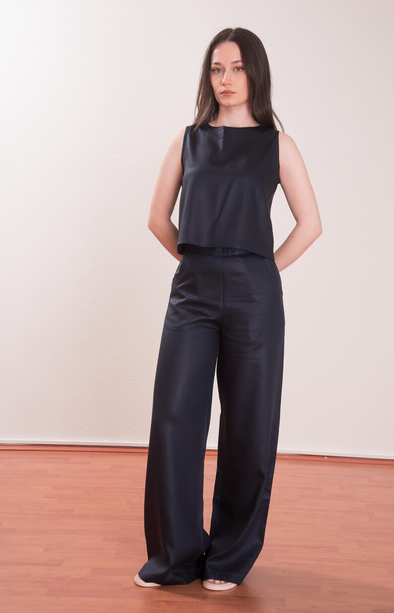 May include: A sleeveless, navy blue top and matching wide-leg pants. The top has a simple, square neckline. The pants are long and flowing. The outfit is made of a smooth, possibly satin-like fabric. The model is standing in front of a plain wall.