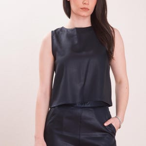 May include: Sleeveless navy blue top and matching pants. The top has a layered design, and the pants appear to be wide-legged. The outfit is made of a shiny material, creating a sleek, modern look. The model is wearing a silver bracelet.