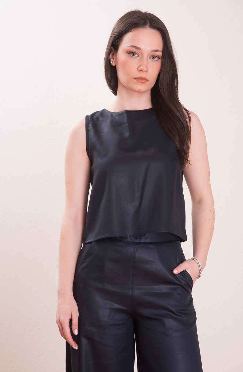 May include: Sleeveless navy blue top and matching pants. The top has a layered design, and the pants appear to be wide-legged. The outfit is made of a shiny material, creating a sleek, modern look. The model is wearing a silver bracelet.