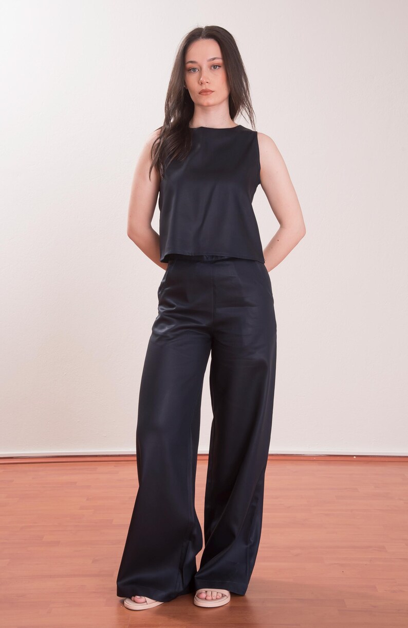 May include: A sleeveless, navy blue top and matching wide-leg pants. The outfit has a sleek, satin-like appearance. The model is standing in front of a plain background and wooden floor.