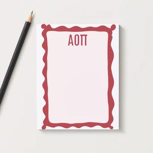 May include: A rectangular notepad with a red wavy border and the letters "AOTT" in the center. A black pencil with a gold ferrule rests on the white surface next to the notepad. The notepad has a light pink interior.