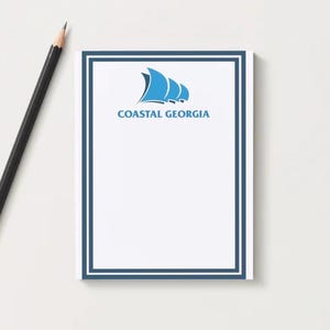 May include: A white notepad with a blue border and the text "COASTAL GEORGIA" and a blue sailboat logo. A black pencil with a gold ferrule and eraser rests on the left side of the notepad.