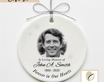 May include: A white ceramic ornament with a black and white photo of a man. The ornament has the text "In Loving Memory of John A. Smith 1950-2025 Forever in Our Hearts." A silver string is attached for hanging. The word "Ceramic" is at the top.