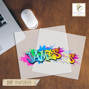 May include: A colorful DTF transfer featuring the name "JAMES" in graffiti style. The name is rendered in vibrant hues of blue, green, yellow, and pink, with a splash effect. The image is on a transparent sheet, with a computer mouse and laptop visible.