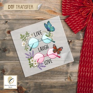 May include: A DTF transfer featuring the words "Live Laugh Love" with round eyeglasses, butterflies, and floral accents. The top glasses have blue lenses, and the bottom glasses have pink lenses. The design is on a light gray background.