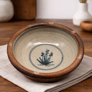 May include: A ceramic bowl with a brown rim and a beige interior. The bowl features a blue floral design in the center. It sits on a gray linen napkin on a wooden surface. Other decorative items are in the background.