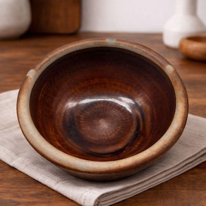 May include: A handcrafted, brown ceramic bowl with a light beige rim, resting on a neutral-colored linen napkin. The bowl's interior has a glossy finish, and the exterior has a textured appearance. The bowl is set on a wooden surface.