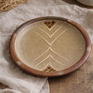 May include: A handmade ceramic plate with a brown rim and a beige center. The plate features a geometric design of white lines and dotted patterns. The plate is displayed on a neutral-colored linen cloth.