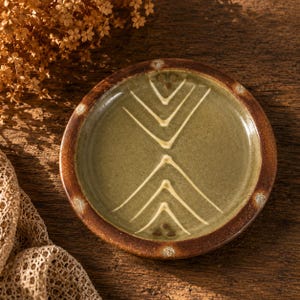 May include: A small, handmade ceramic plate with a brown rim and a green center. The plate features a white geometric design. The plate is approximately 5 inches in diameter.