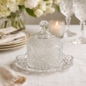 May include: A clear, decorative glass butter dish with a diamond pattern and a crystal knob on top. The dish sits on a matching scalloped plate. The background includes white flowers, candles, and glassware, suggesting a formal dining setting.