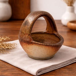May include: A handmade ceramic basket with a handle. The basket is a warm, earthy tone with brown and tan hues. It sits on a neutral-colored linen napkin, suggesting a rustic or farmhouse aesthetic.