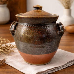 May include: A handcrafted ceramic pot with a lid, featuring a dark brown and reddish-orange color scheme. The pot has two handles and is placed on a neutral-colored cloth. The rustic design suggests it's suitable for kitchen or home decor.