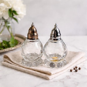 May include: Two clear glass salt and pepper shakers with textured, round bodies and decorative metal lids. The shakers sit on a beige linen napkin. One lid is bronze, the other is silver. A few peppercorns are scattered nearby.