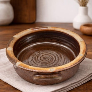 May include: A brown ceramic baking dish with a spiral pattern in the center. The dish has a rounded shape with a wide rim and small handles. It sits on a beige linen napkin on a wooden surface. The rim has a lighter, tan color.