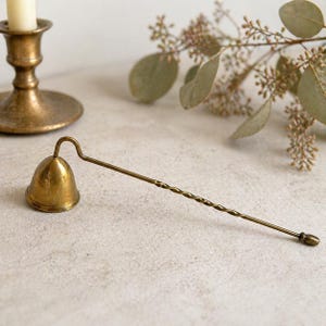 May include: A vintage brass candle snuffer with a bell-shaped head and a long, twisted handle. The snuffer is designed to extinguish candles and is placed on a neutral surface with a brass candlestick in the background.