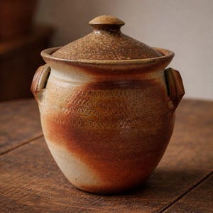 May include: A handcrafted ceramic jar with a lid. The jar is a warm brown color with a textured surface and a diagonal gradient of orange and cream. It has two small handles and sits on a wooden surface.