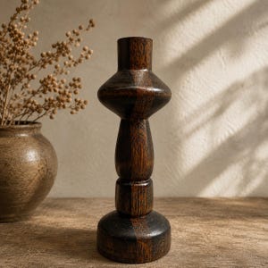 May include: A dark brown wooden candlestick holder with a unique, layered design. The holder has a wide, circular base, a cylindrical midsection, and a flared top. The wood grain is visible, adding texture. A vase with dried flowers is in the background.