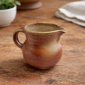 May include: A small, handmade ceramic creamer with a rounded body and a curved handle. The creamer is glazed in shades of brown and tan, with a speckled texture. It has a spout for easy pouring and is likely used for serving cream or milk.