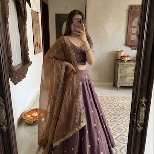 May include: A traditional Indian outfit featuring a purple skirt with white dot accents, a matching embroidered top, and a sheer brown shawl. The outfit is reflected in a mirror, showcasing the intricate details and design.