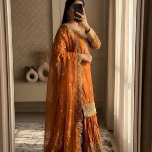 May include: An orange sharara suit with gold embroidery. The outfit includes a long-sleeved top, wide-legged pants, and a sheer dupatta. The suit has intricate gold detailing around the neckline, cuffs, and hem. The brand name "SAANJHHNA" is visible at the top.