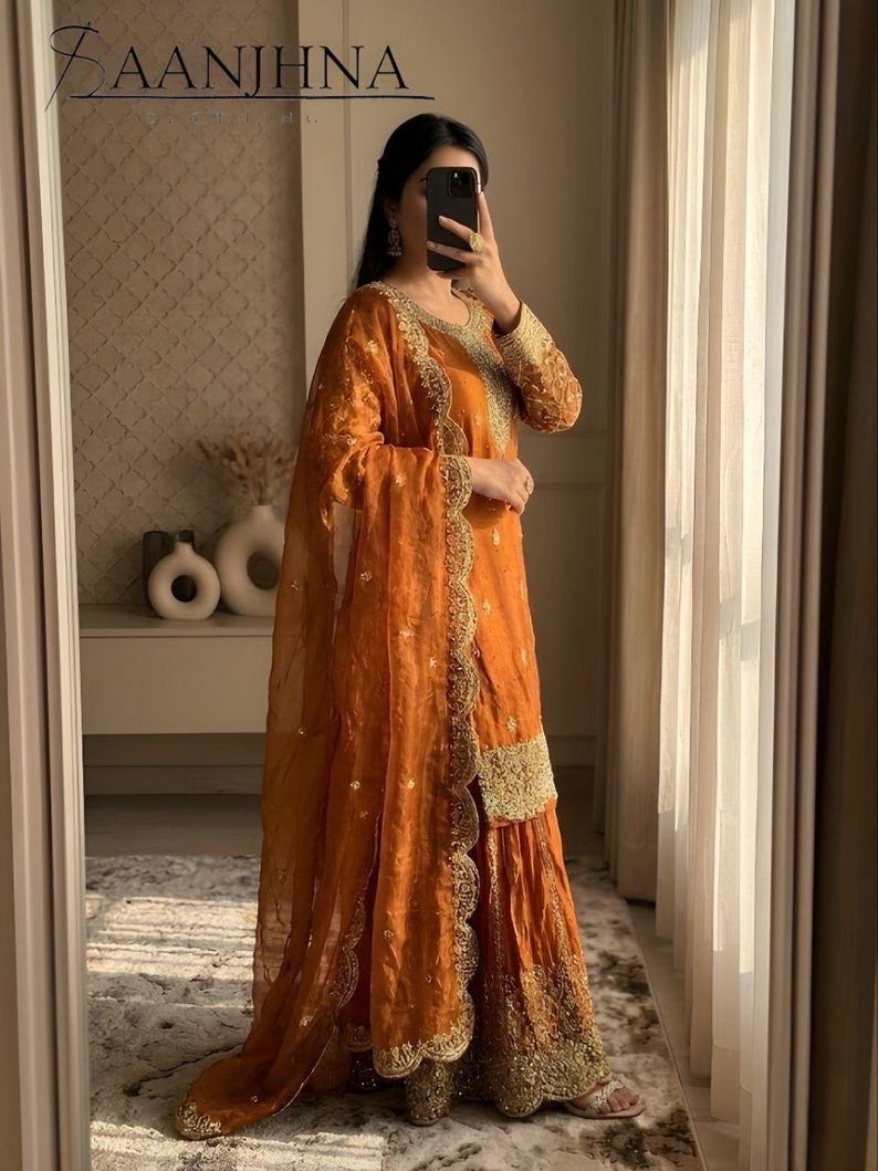 May include: An orange sharara suit with gold embroidery. The outfit includes a long-sleeved top, wide-legged pants, and a sheer dupatta. The suit has intricate gold detailing around the neckline, cuffs, and hem. The brand name "SAANJHHNA" is visible at the top.