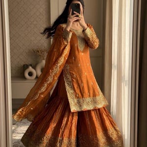 May include: Orange Sharara suit with gold embroidery. The outfit includes a long-sleeved tunic and wide-legged pants. The outfit is made of a shiny fabric and has gold trim along the edges. The outfit is worn with a matching dupatta.
