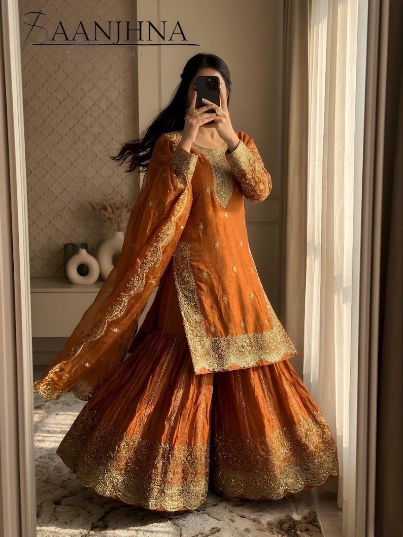 May include: Orange Sharara suit with gold embroidery. The outfit includes a long-sleeved tunic and wide-legged pants. The outfit is made of a shiny fabric and has gold trim along the edges. The outfit is worn with a matching dupatta.