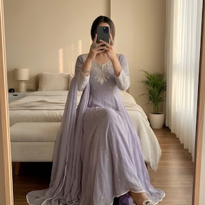 Peut inclure: Une robe longue lilas avec des broderies complexes sur le corsage et les manches. La robe présente une silhouette fluide et est associée à une écharpe assortie. La tenue est complétée par des chaussures ornées.