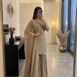 May include: A beige embroidered sharara suit with a matching dupatta. The outfit features intricate detailing and embellishments. The image includes the text "SAANJNA CLOTHING" at the top. The outfit is displayed in a well-lit room with a large mirror.
