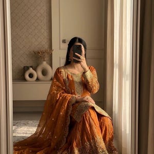 May include: An orange salwar kameez set with gold embroidery. The traditional outfit includes a long-sleeved top, wide-legged pants, and a sheer dupatta. The outfit is worn by a person in a mirror reflection.