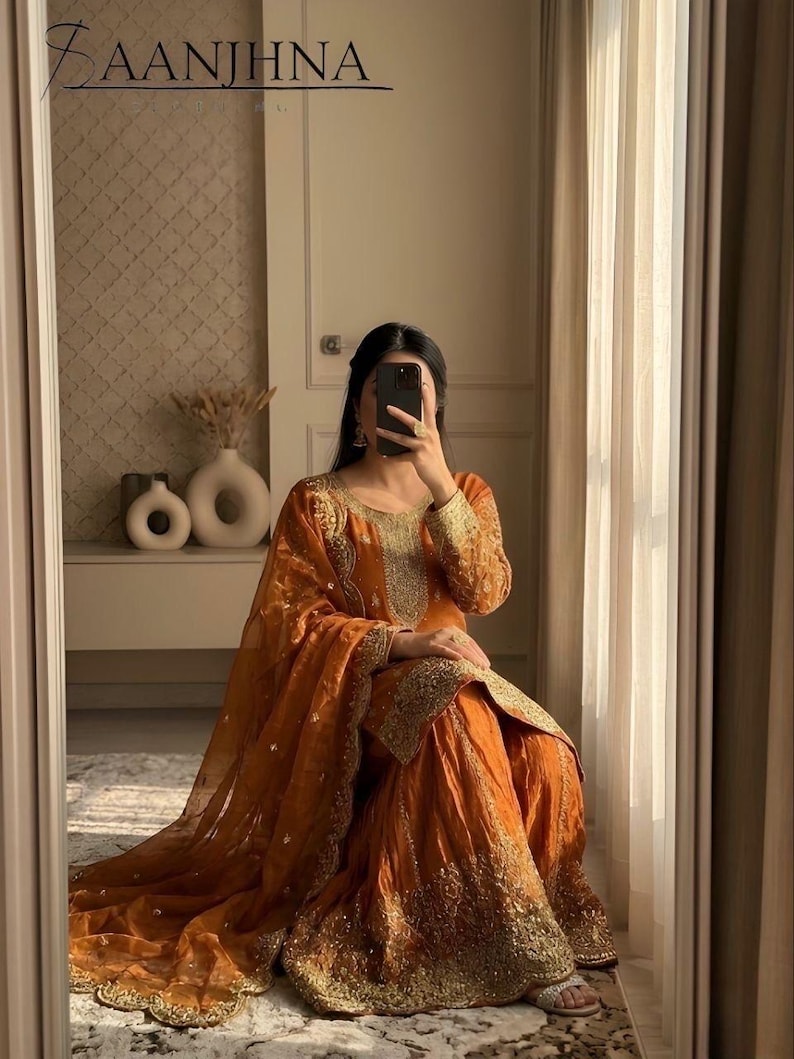 May include: An orange salwar kameez set with gold embroidery. The traditional outfit includes a long-sleeved top, wide-legged pants, and a sheer dupatta. The outfit is worn by a person in a mirror reflection.