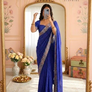 Könnte beinhalten: Ein leuchtend blaues Saree mit Streifenmuster und aufwendigem Rand. Das Saree wird mit einer passenden Bluse kombiniert. Das Bild zeigt auch einen goldgerahmten Spiegel und den Text "SAANJHIINA CLOTHING".