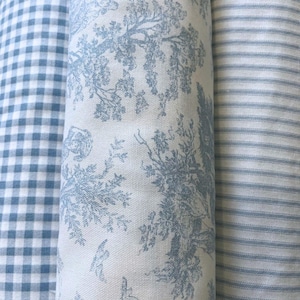 May include: Three fabric rolls with different patterns. One is a blue and white gingham check, another features a blue toile design with trees and figures, and the third has blue and white stripes. The fabrics are tightly rolled.