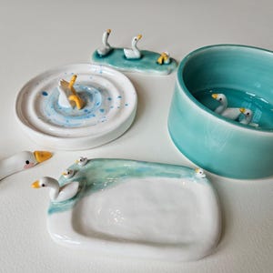 May include: A collection of ceramic items featuring white geese with yellow beaks. The set includes a round dish with a goose, a rectangular tray with small geese, a small bowl with two geese inside, a long tray with two geese, and a single goose figurine.