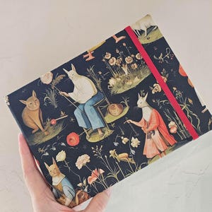 May include: A rectangular notebook with a black cover featuring a repeating pattern of anthropomorphic cats in historical clothing, flowers, and foliage. The notebook is secured with a bright red elastic band that stretches across the front cover.