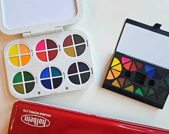 Könnte beinhalten: Eine weiß gesprenkelte Kunststoffpalette mit sechs kreisförmigen Vertiefungen, die vierteilige Farbsegmente enthalten, eine schwarze rechteckige Palette mit dreieckigen Farbsegmenten und ein großer roter Metallkasten von Holbein Works, Ltd.