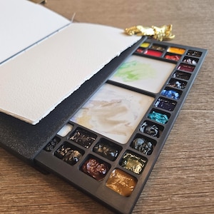 May include: A black portable watercolor palette set featuring a rectangular case with numerous small paint pans in various colors, including red, blue, green, and brown, alongside a white mixing area and a stack of blank white watercolor paper.