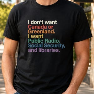 May include: A black short-sleeved t-shirt featuring multi-colored text that reads: I don't want Canada or Greenland. I want Public Radio, Social Security, and libraries. The shirt has a crew neckline and is worn with blue denim jeans.