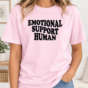 May include: A light pink short-sleeved t-shirt featuring the black text "EMOTIONAL SUPPORT HUMAN" printed in a bold, wavy, retro-style font across the center of the chest. The garment has a classic crew neckline and a standard straight-cut fit.