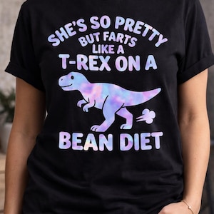 May include: Black t-shirt with the text "SHE'S SO PRETTY BUT FARTS LIKE A T-REX ON A BEAN DIET" in pastel colours. A cartoon dinosaur in similar colours is also featured on the shirt.